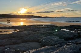 A scenic photo of the Gulf of Maine shoreline showing two people walking into a sunset.